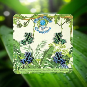 May include: A card featuring four pairs of frog-shaped earrings. Two pairs are green with black spots, and two are blue with black spots. The card has a jungle theme with green leaves and vines. The text "Moon Shark Creations" is visible.
