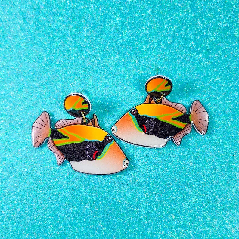 Handmade rectangular triggerfish earrings crafted from shrink plastic with a glossy UV resin front. These lightweight dangle earrings feature bold rectangular triggerfish with striking black bodies, bright yellow markings, and electric blue accents inspired by tropical reef life. Eye-catching and unique, they add a vibrant ocean vibe—perfect for marine life lovers and statement jewelry fans. By Moon Shark Creations.