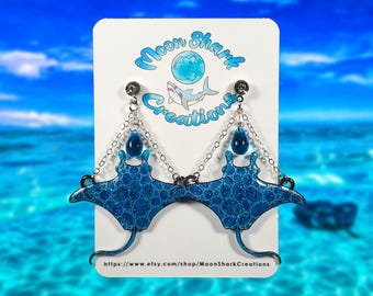 May include: Blue manta ray earrings with a textured pattern and silver chain accents. The earrings are attached to a white card with the text "Moon Shark Creations" and a shark graphic. The background is a blurred image of the ocean.