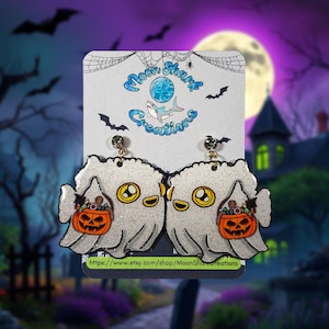 May include: Glittery ghost earrings with a Halloween theme. Each earring features a cute ghost holding a pumpkin-shaped candy bucket. The earrings have a gold-colored stud and are on a backing card with the text "Moon Shark Creations".