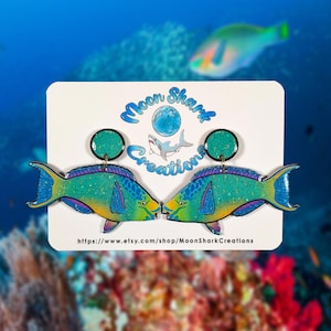 Könnte beinhalten: Ein Paar farbenfrohe Fischohrringe in Türkis, Gelb und Lila. Die Ohrringe sind auf einer weißen Karte mit dem Text "Moon Shark Creations" und einer Website-Adresse abgebildet. Der Hintergrund zeigt eine unscharfe Unterwasserszene mit einem Fisch.