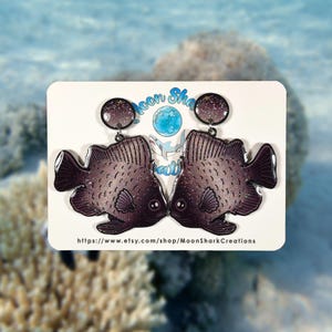 May include: A pair of fish-shaped earrings with a dark purple and glittery design. The earrings are displayed on a white card with the text "Moon Shark Creations" and a website address. The background shows an ocean scene.