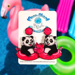 May include: A pair of novelty earrings featuring two pandas and a pig in a red inflatable ring. The earrings have a red glitter circle at the top and are set against a background of pink and blue pool floats. The text "Moon Shark Creations" is visible.