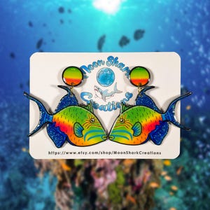 May include: A pair of colorful fish-shaped earrings. The earrings feature a vibrant design with green, yellow, orange, and blue hues. The earrings are displayed on a white card with the text "Moon Shark Creations" and a website address.
