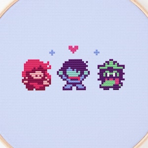 May include: A cross-stitch embroidery hoop featuring three pixelated characters in red, purple, and green, with a pink heart and blue crosses. The design is set against a light blue fabric, with the text "@chai stitch cafe" at the bottom.