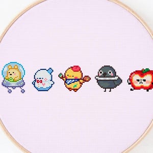Puede incluir: Un aro de bordado rosa con fondo blanco. Cinco personajes pixelados están bordados en el aro. Los personajes son un pollito amarillo con un pincel, un fantasma blanco, un perro marrón en una nave espacial, un pájaro negro y una manzana roja con cara.