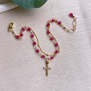 May include: A layered necklace with a gold chain and a cross pendant. The necklace features small, round, red beads. The cross pendant is gold and has a textured design. The necklace is on a cream-colored surface.