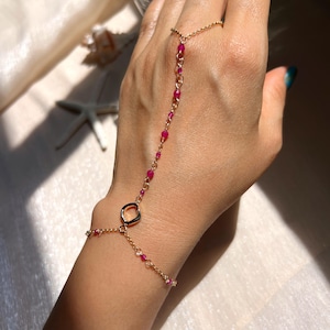 Puede incluir: Una cadena de mano dorada con un anillo, una pulsera y una cadena para el dedo, adornada con pequeñas cuentas rojas vibrantes. La joya está diseñada para ser usada en la mano, conectando el anillo a la pulsera con una delicada cadena.