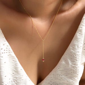 Puede incluir: Un delicado collar dorado con un diseño en forma de Y. El collar presenta una serie de pequeñas cuentas a lo largo de la cadena y un grupo de cuentas rojas al final de la caída. El collar se usa sobre una blusa blanca.