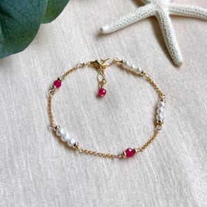 Op de afbeelding: Een delicate gouden kettingarmband met afwisselend witte parels en rode kralen. De armband heeft een kleine, hangende rode kraal en een gouden sluiting. Een witte zeester staat op de achtergrond.