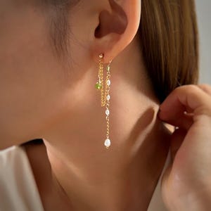 Peut inclure: Boucles d'oreilles pendantes dorées avec un design de chaîne délicate. Les boucles d'oreilles présentent de petits accents verts et blancs, dont des perles. Les boucles d'oreilles sont portées sur une oreille, la chaîne tombant.