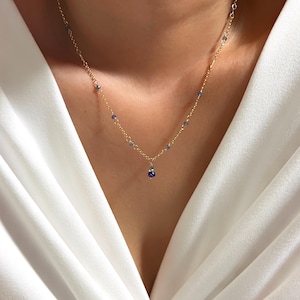 Può includere: Delicata collana d'oro con piccole perline blu e un pendente blu rotondo più grande. La collana è indossata su uno sfondo di tessuto bianco, evidenziando il design elegante del gioiello.