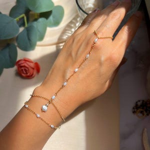 Könnte beinhalten: Eine zarte goldene Handkette mit kleinen, weißen Perlen. Die Kette verbindet sich mit einem Ring und umschließt das Handgelenk, wodurch ein mehrschichtiger Armbandeffekt entsteht. Der Schmuck wird auf einer Hand präsentiert.