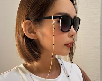 Könnte beinhalten: Eine goldfarbene Brillenketten mit schwarzen Perlen ist an einer schwarzen Sonnenbrille befestigt. Die Kette hängt herunter, und die Sonnenbrille wird von einer Person getragen. Die Person trägt ein weißes Oberteil.