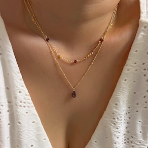Puede incluir: Un collar dorado en capas con un colgante de lágrima. El collar presenta dos hilos, uno con pequeñas cuentas rojas y amarillas, y el otro con un colgante de piedra preciosa roja oscura en forma de lágrima. El collar se usa sobre un fondo blanco.