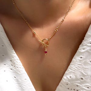 Könnte beinhalten: Eine goldene Kettenkette mit einem Toggle-Verschluss und einem einzelnen roten Edelstein-Anhänger.