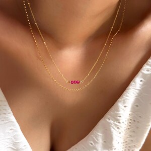 Op de afbeelding: Een goudkleurige ketting met twee strengen en fijne kettingen. De onderste ketting heeft drie kleine, helder roze edelstenen. De kettingen worden gedragen over een witte top.