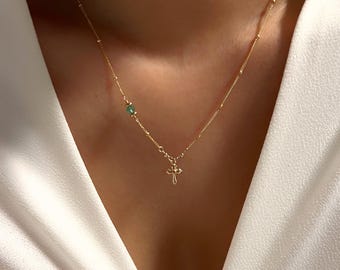 May include: Delicate gold necklace featuring a small cross pendant and a single green gemstone. The necklace has a fine chain with small, evenly spaced beads. The cross pendant is intricately detailed. The necklace is displayed against a white background.
