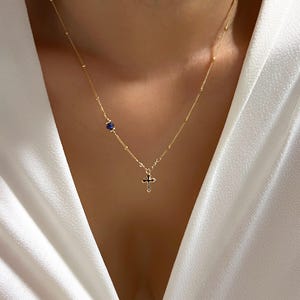 Op de afbeelding: Een gouden ketting met een klein blauw edelsteentje en een zwarte kruis hanger.