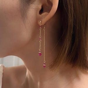 Könnte beinhalten: Ein Paar lange, goldene Kettenohrringe mit kleinen roten Edelstein-Akzenten. Die Ohrringe hängen vom Ohrläppchen herab und haben an jedem Kettenende einen größeren roten Edelstein.