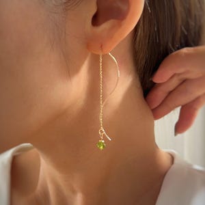May include: A gold-toned threader earring featuring a long, curved, textured wire that loops through the earlobe. A small, faceted, lime-green gemstone bead hangs from the bottom of the curved wire, creating a delicate and dangling silhouette.