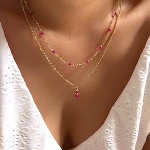 May include: A layered gold necklace with small, vibrant pink gemstones. The necklace features two strands, one with evenly spaced gems and the other with a pendant of three gems. The jewelry is displayed against a white background.