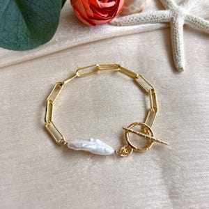 May include: Gold-toned chain bracelet featuring a large, irregular white pearl. The bracelet has a toggle clasp and a series of rectangular links. The background is a light, neutral color.