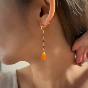 May include: A gold hoop earring featuring a delicate chain drop. The chain is accented with three small, round, deep red beads and terminates in a single, larger, teardrop-shaped orange stone pendant, all connected by thin gold-toned metal links.