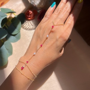 May include: A delicate gold hand chain bracelet with a ring, adorned with small red and white beads. The jewelry piece is worn on a hand with painted nails, showcasing the bracelet's design and details.