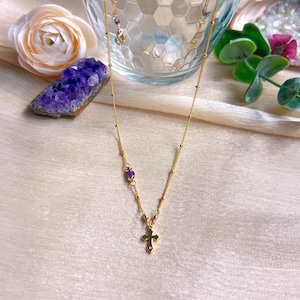 May include: A delicate gold necklace featuring a cross pendant with a small gemstone. The necklace also includes small purple gemstones and a beaded chain. The background includes a purple crystal and a rose.
