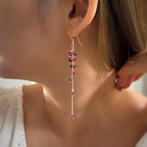 May include: Gold-tone dangle earrings with a cascade of colorful gemstones. The earrings feature a cluster of red, purple, and pink beads, suspended from a delicate chain. The earrings are designed to be worn in pierced ears.