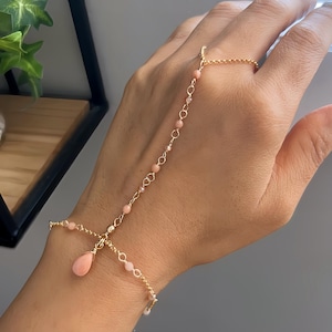 Könnte beinhalten: Ein goldenes Kettenarmband mit rosa Perlen und einem rosa Tropfen-Anhänger. Das Armband ist so konzipiert, dass es am Handgelenk und am Finger getragen werden kann.