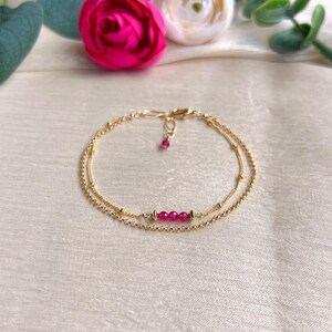 May include: A delicate gold bracelet with two strands. The top strand has a small ruby pendant, and the bottom strand features a row of three rubies. The bracelet is set against a soft, cream-colored background with pink flowers.