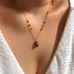 May include: A delicate gold necklace featuring a teardrop-shaped tiger's eye pendant. The necklace has small, round, brown beads and a fine gold chain. The pendant is a warm brown color with golden streaks.