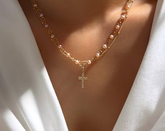 May include: A gold chain necklace with a small gold cross pendant. The chain is layered with a strand of small pink pearls.