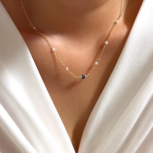 Puede incluir: Delicado collar de oro con pequeñas perlas blancas y una sola gema azul oscuro. El collar se muestra sobre un fondo blanco, resaltando el elegante diseño. Una fina pieza de joyería.