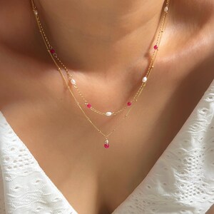 May include: A delicate gold necklace with two strands. The top strand features small white pearls and red beads, while the bottom strand has a single red pendant. The necklace is layered and sits on a white top.