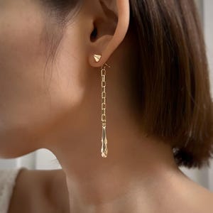 Puede incluir: Pendientes de cadena de oro con un pequeño triángulo en la parte superior y un dije en forma de lágrima en la parte inferior.