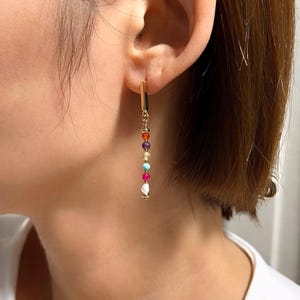 May include: Gold-colored dangle earrings with a vertical bar design. The earrings feature a series of colorful beads in various shades, including red, orange, purple, yellow, turquoise, pink, and white. The earrings are designed to be worn in pierced ears.
