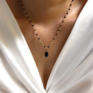Puede incluir: Un delicado collar dorado con pequeñas cuentas negras y un colgante negro en forma de lágrima. El collar se lleva sobre una blusa blanca, resaltando el contraste entre el oro y el negro.