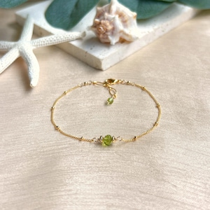 May include: A delicate gold chain bracelet featuring a single small, round, translucent green peridot bead centred between two tiny gold beads. The bracelet includes a thin gold chain with evenly spaced gold accents and a lobster claw clasp.