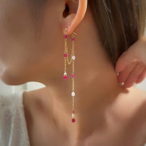 Op de afbeelding: Goudkleurige oorhangers met een kettingontwerp. De oorbellen hebben afwisselend roze en witte kralen. De oorbellen worden getoond op het oor van een persoon.