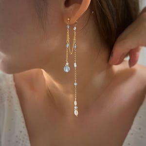 May include: Gold-toned dangle earrings with delicate chains and light blue and white beads. The earrings feature a small, round, light blue crystal at the base of one chain and a pearl at the end of the other.