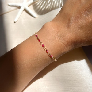 Könnte beinhalten: Ein zartes goldenes Armband mit kleinen, leuchtend roten Perlen. Das Armband ist an einer feinen Kette aufgereiht und wird an einem Handgelenk präsentiert. Der Schmuck ist schlicht und elegant, geeignet für den Alltag oder besondere Anlässe.