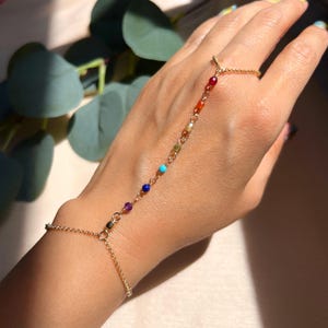 May include: A gold hand chain bracelet featuring a series of colourful beads. The beads are in a rainbow spectrum of colours, including red, orange, yellow, green, blue, and purple. The bracelet connects to a ring and a wrist chain.
