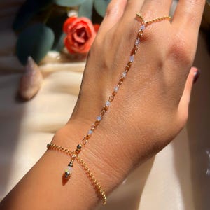 Könnte beinhalten: Eine zarte goldene Handkette mit einem Ring und einem Armband, die durch eine Kette verbunden sind. Die Kette hat kleine, schillernde Perlen und einen hängenden Anhänger. Der Schmuck wird auf einer Hand präsentiert und zeigt sein kompliziertes Design und seinen eleganten Stil.