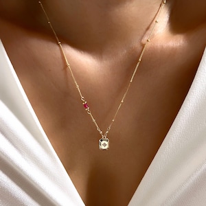 May include: A gold chain necklace with a small square pendant featuring a star design. The chain has small red beads.