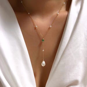 Peut inclure: Un collier en chaîne dorée avec une grosse perle pendante et un petit accent de pierre précieuse verte.