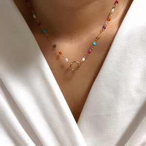 Könnte beinhalten: Eine goldfarbene Halskette mit einem kreisförmigen Anhänger. Die Kette ist mit bunten Perlen verziert, darunter rosa, türkis, orange und weiß. Die Halskette wird vor einem weißen Kleidungsstück getragen.