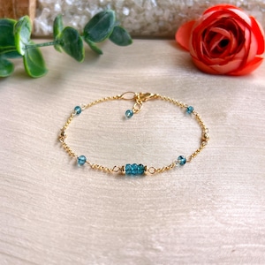 Puede incluir: Una delicada pulsera dorada con pequeñas cuentas redondas de color azul claro y un grupo de tres cuentas azules más grandes. La pulsera tiene un diseño de cadena simple con un cierre de broche. La joya se exhibe sobre una superficie de color claro.